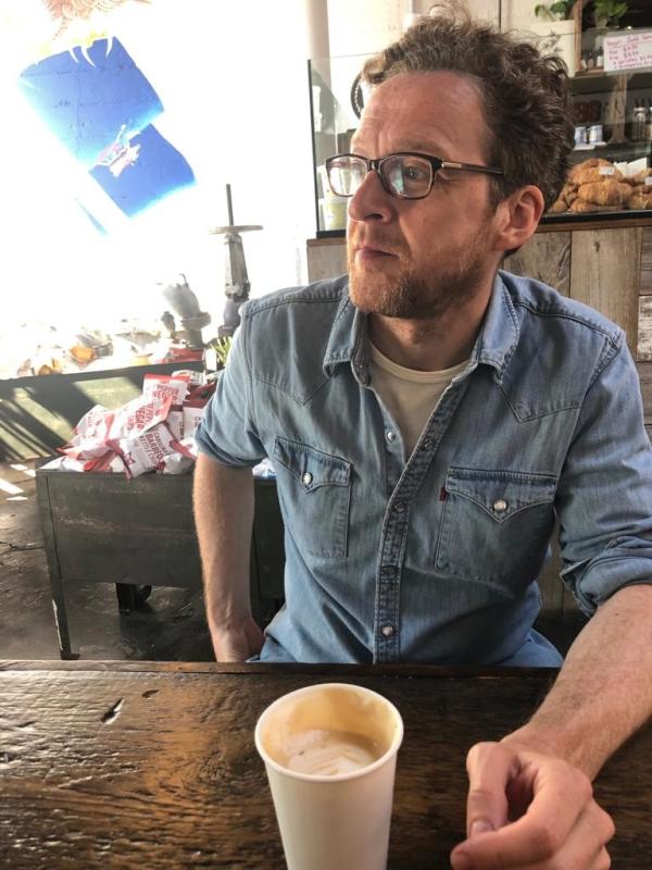 A man with a bear, glasses and brown hair looking to his right seated at a wooden table with a cup of coffee in front of him