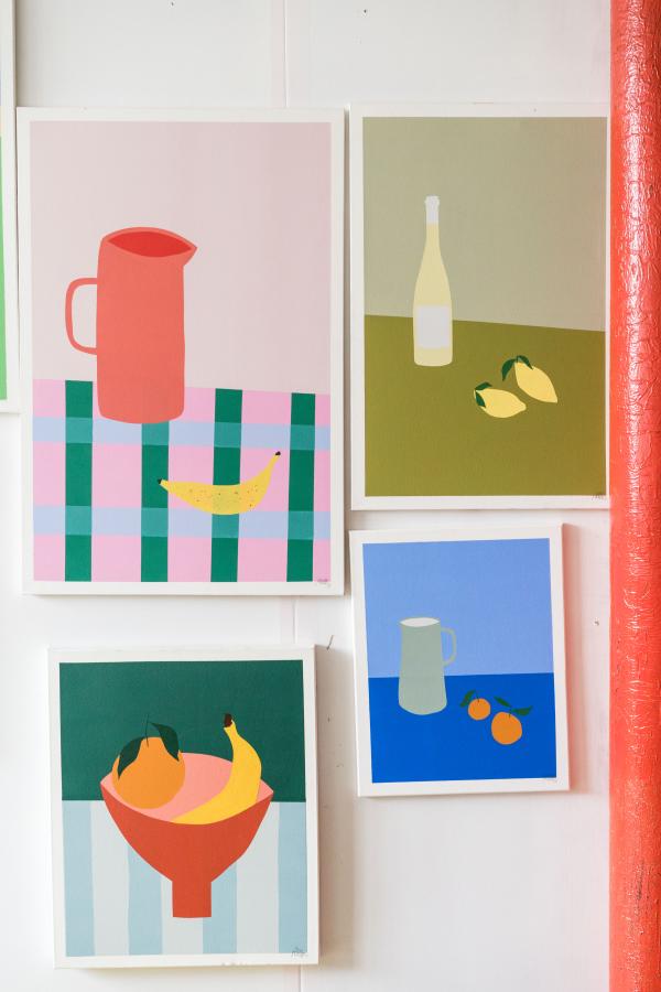 4 paintings hang on a white wall, each featuring colourful, stylised still lives.