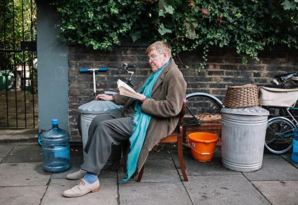 A photograph featuring Alan Bennett sat on a chair outside reading a book.