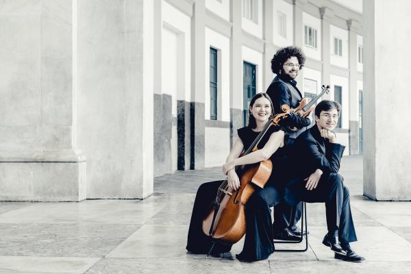 In the background are lots of angular white marble buildings with blue/green doors. There are three people in the foreground, two are white men and one white woman. One of the men has a mass of dark curly hair, glasses, a beard and is holding a violin. The other man is sat cross legged with his elbow on his knee and arm bend up towards his face. He is has shorter light brown hair and is also smiling. The third person is a white woman with long dark dark, and has her arms wrapped around a cello.