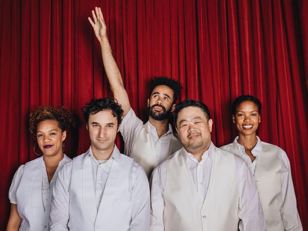 Five people wearing white shirts and suits stood in front of a red curtain. The man in the middle of the image has one arm in the air.