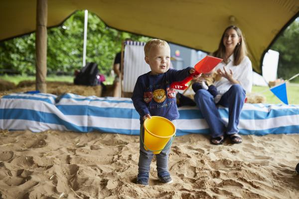 A young blonde child playing in a covered outdoor sand pit. A woman with brown mid-length hair looks on, laughing.