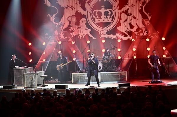 Queen Extravaganza band members performing on stage in front of a red backdrop