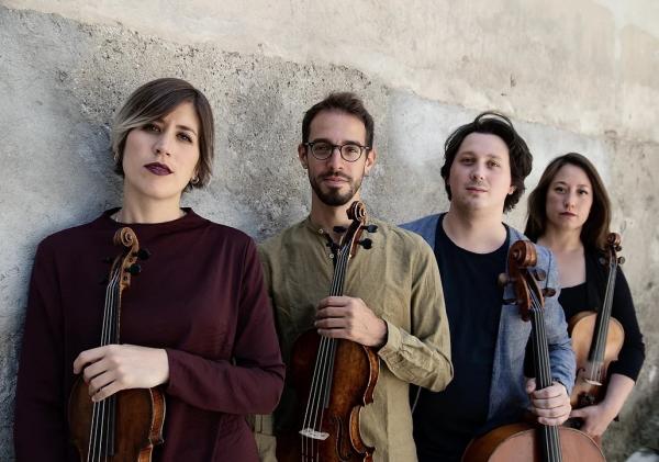 There are four people in this image, of Italian heritage. The first is a white woman with short brown to ash blonde ombre hair. She's wearing a dark red top and has dark features and lipstick, and is holding a violin. The second is a man wearing glasses with a beard, wearing a muted olive green top. He's holding a violin. The third person is a man with longer floppy hair, wearing a black tshirt with denim blue over shirt. The fourth person is a woman with long dark hair and a black top she's holding a viola