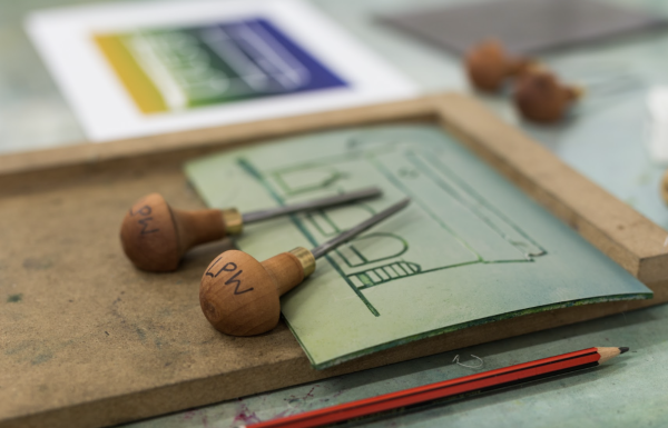 The image shows a piece of lino on a wooden board with linocutting tools.