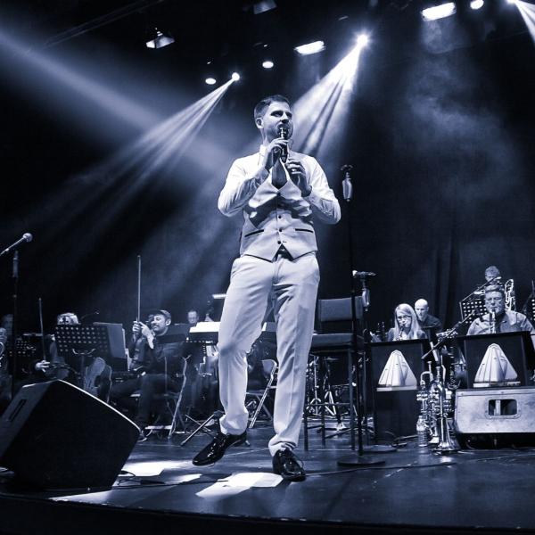 Grey-toned image of a short-haired man holding a microphone to his mouth in a waistcoat and tie with the rest of the swing band in the background.