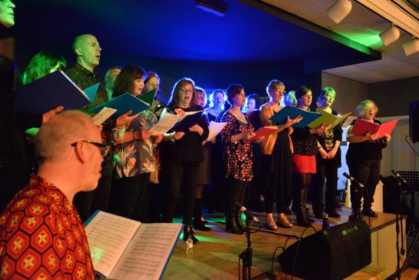 A choir of men and women are stood on a stage singing together holding music folders.