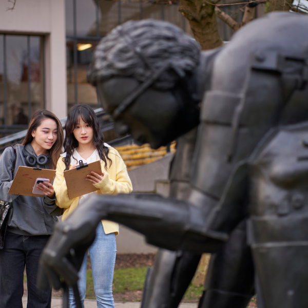 Two people with clipboards stood drawing a bronze sculpture of a man bent over in thought