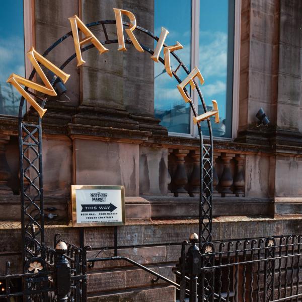 Image shows outside of The Northern Market, with a metal archway that reads "MARKET" in large gold letters and a sign saying the venue is down the stairs.
