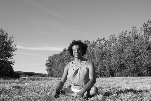 Asher sat in a field, clear sky, trees in background. He is facing and smiling at the camera, dressed for yoga.