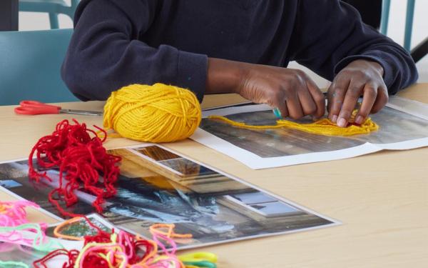 A pair of hands use yellow wool to trace an image on a page. There's a jumble of wool and images also on the table