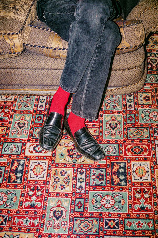 A person sits cross-legged on a patterned couch, wearing black jeans, bright red socks, and black leather loafers. The colorful carpet beneath features a detailed, intricate pattern of red, blue, green, and beige. The shot focuses on the lower half of the body, highlighting the contrast between the red socks and the rest of the muted tones.