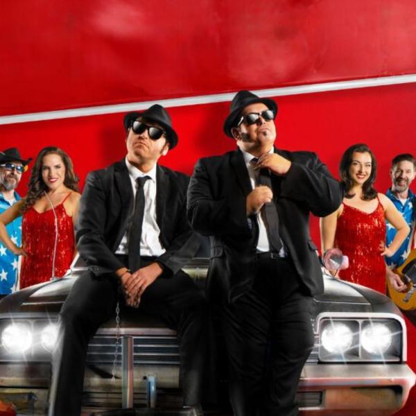 Two men in black suits sat on the bonnet of an old looking car. Behind the car in an arrow-like formation are men wearing American flag shirts and two women in red fringe dresses appear with the band.