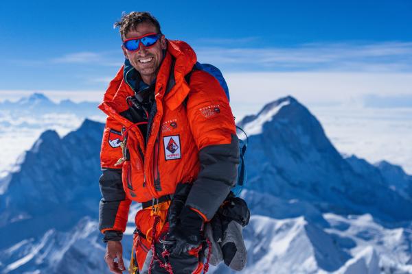 A man in an orange snow suit and sunglasses, with a snowy mountain peak in the background.