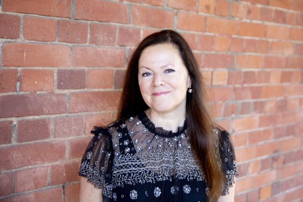 A white woman in her mid 60s. She has long straight dark hair, she is smiling, and is wearing a black sheer top with white details. There is a red brick wall behind her. 