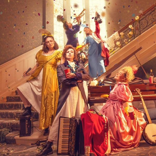 A group of people in period dress on and next to the bottom of a staircase holding various more modern items such as a microphone and red solo cups as well as instruments such as a banjo, a piano and a trumpet also appearing. Confetti is falling from the ceiling. 