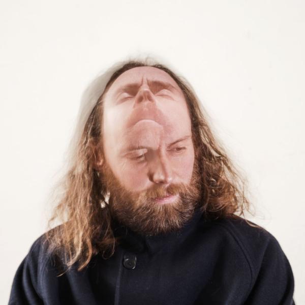 Man with long hair and a beard in a black top looking slightly down with an image of him looking up merged into the top of his head.