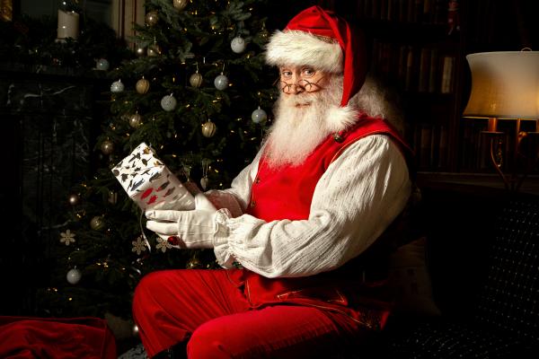 Santa Claus sits in front of a Christmas Tree holding a wrapped Christmas present