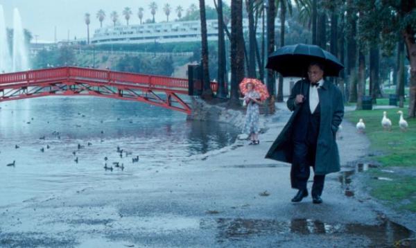 A man in a tuxedo walks next to a red bridge holding an umbrella.