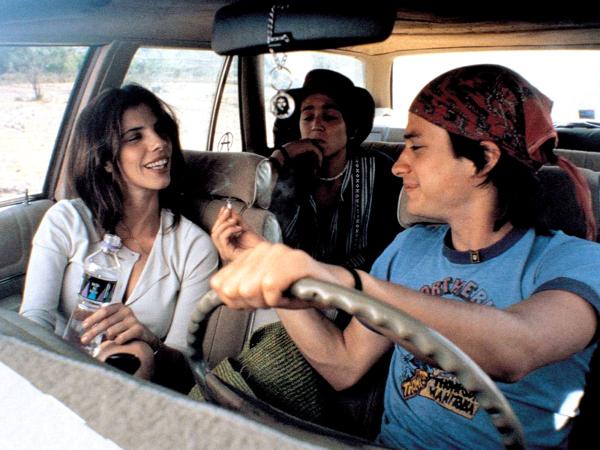 A young man, in the driver's seat of a car, passes a cigarette to the older woman next to him, while anther young man sits behind.