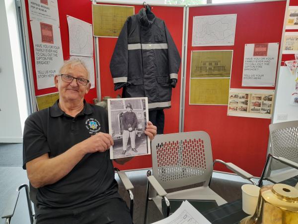 A white, older man sits on a chair, holding a black and white photography of a fireman. In the background there is a firesuit hanging on a board with other old artifacts. 