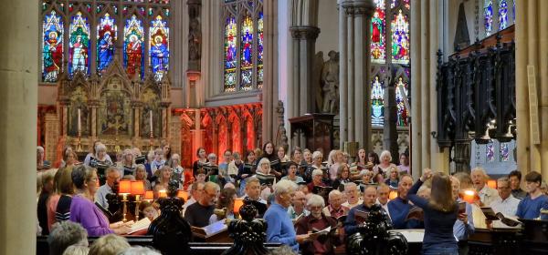 A group of singers holding music scores stand singing in a church. A conductor is standing with her back to the camera, arms raised