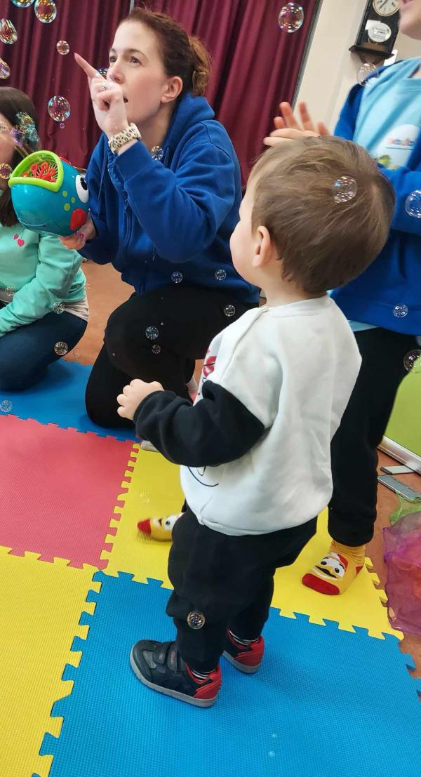 An adult with a bubble machine blowing bubbles a toddler with his back to the camera watching the bubbles.