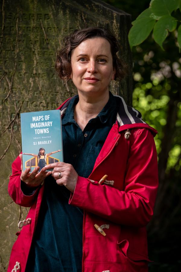 Photo of a woman in a hot pink coat standing by a tree, holding a book with a blue cover titled 'maps of imaginary towns; 