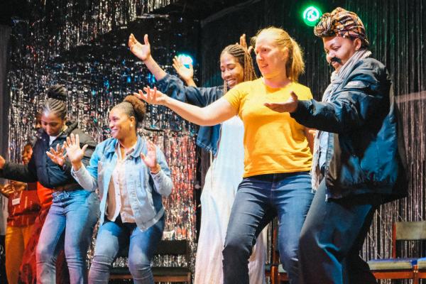 5 women dance together on a stage, they are facing towards an audience, there is a tinsel curtain behind them