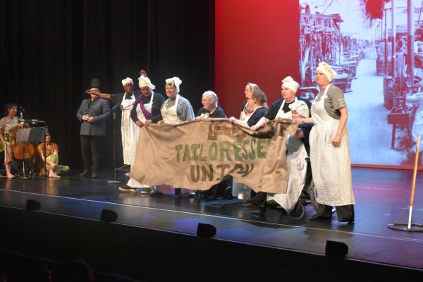 8 women in aprons and hair wraps holding a banner on stage with a man standing in the background. 