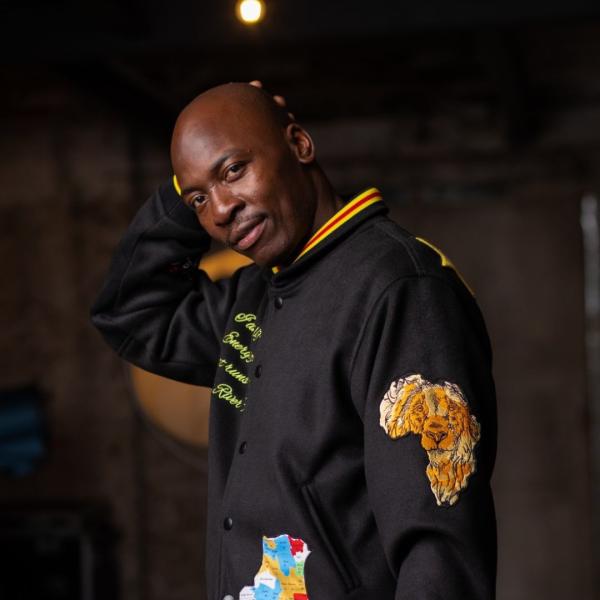 Eddie Kadi wearing a black jacket that has the shape of the continent of Africa featuring a lion's face on his left arm with his right hand behind his head.