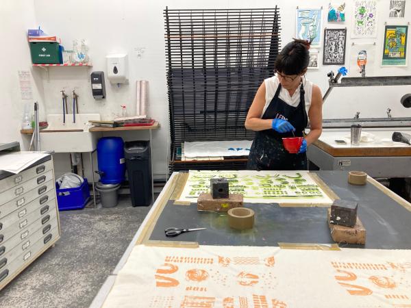 Image shows an artist working in the Leeds Print Workshop space, holding a pot of ink and looking down at their work