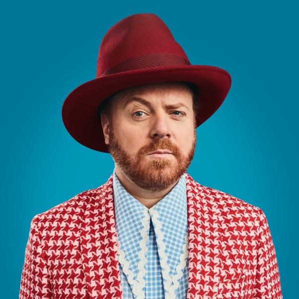 Leigh Francis in a red and white gingham-patterned suit, pale blue and white shirt with long collar, maroon oversized bowler hat. He's raising his right eyebrow.