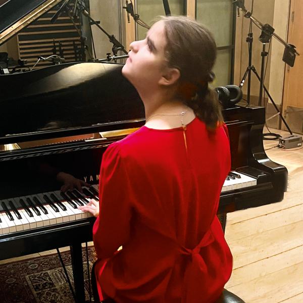 Lucy wearing red sat at a grand piano in a rehearsal studio.