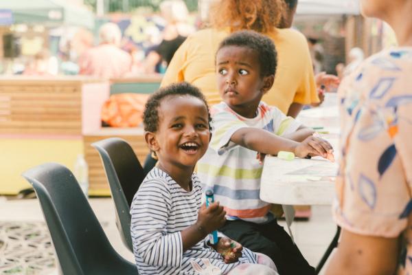 Two young boys are sat outside in front of a white table. One boy is looking at something out of frame, the boy closest to the camera is looking towards us, laughing and holding a coloured pen 