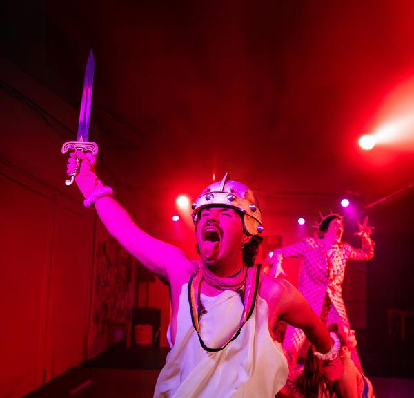  Person in a theatrical performance, wearing a spiked helmet and holding a sword, with another person in the background. The scene is lit with dramatic pink lighting.