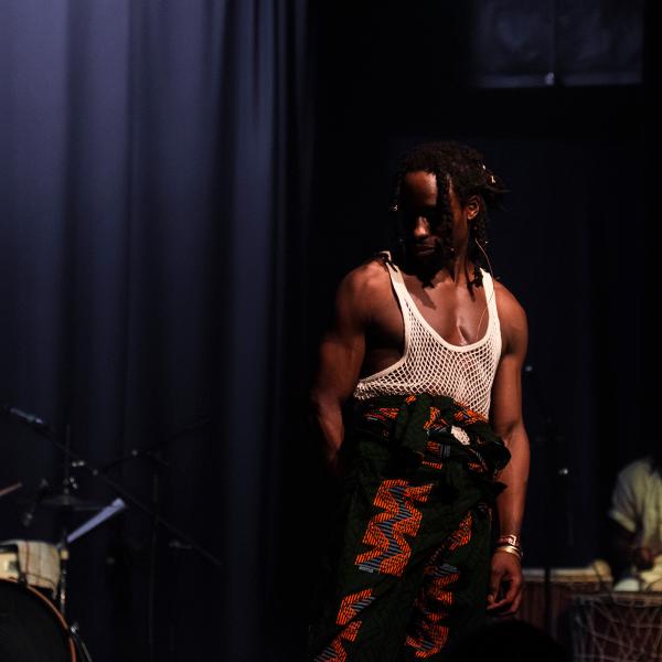 A black man wearing a white string vest standing on stage in front of a band of drummers