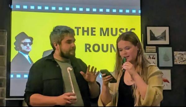 Scalarama Leeds team members playing the kazoo and another fun percussion instrument in the music round of last year's quiz with Blues Brothers on the screen 