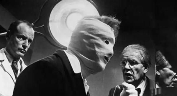 Still from Seconds, bright light is over a man with a bandage all over his face and two other people looking at him one with a doctors style coat on 