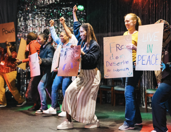 A diverse group of defiant women stand on a stage holding protest signs with messages of peace and freedom.