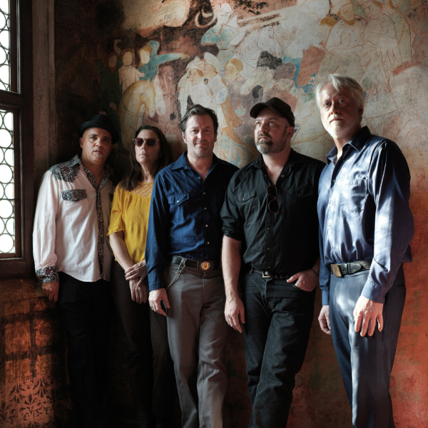 The five members of The Delines stood in different coloured shirts against a beige slightly-patterned wall either looking at the camera or looking in a different direction.