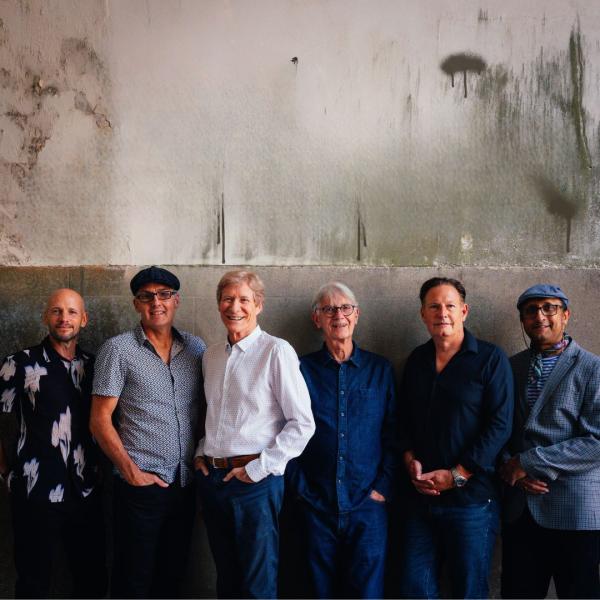 Six middle-aged men in varying blue shirts and jeans stood against a concrete wall.