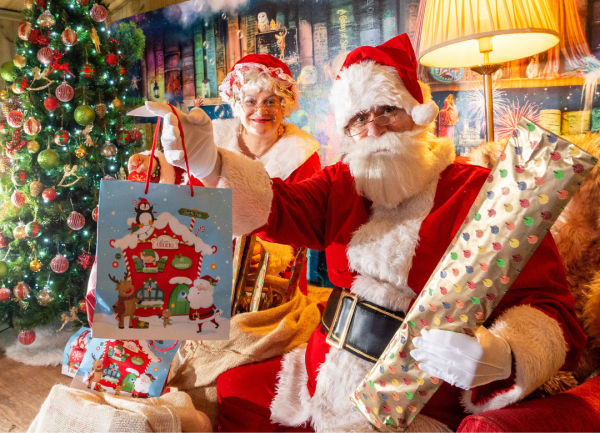 Santa and Mrs Claus sat in a wood cabin next to a decorated Christmas tree. Santa holding two presents and smiling.