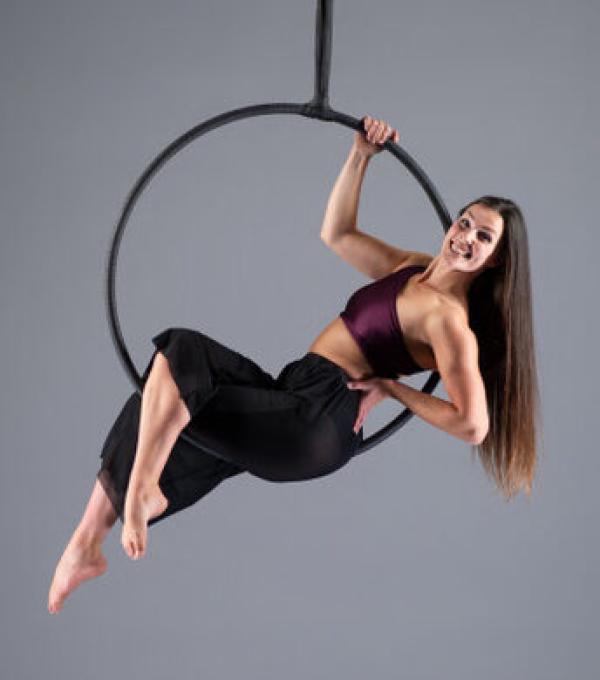 Studio photo of workshop facilitator Jess, sat and reclining back in a hanging hoop. Striking a strong pose and smiling at the camera