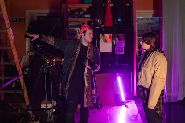 A white man with a camera and red hat points to the left, there is also a white girl with dark hair and beige jacket, a bring pink neon light can be seen on the floor.