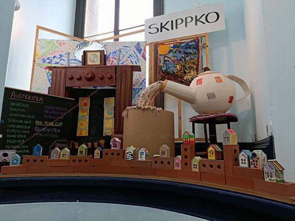 Artworks on display at Leeds City Museum including a large cardboard fireplace and teapot, a handmade rag rug showing the sun and a tree. In the foreground there is a row of house bricks with small decorated wooden houses standing on top.