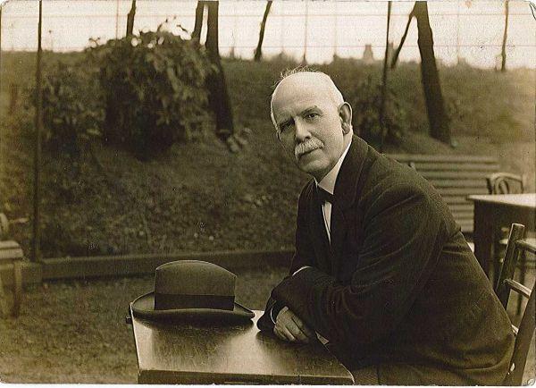 A sepia photo of Dr James Allan sat outside at a table. He is smartly dressed and his hat is left on the table.