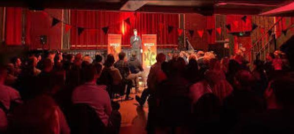 A person with a microphone stands on a stage in a red lit room, performing to a large crowd.