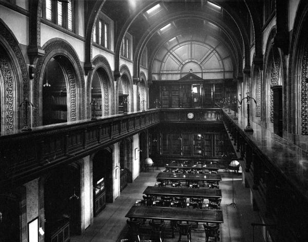 Black and white photo looking from gallery to the main reference library. Long tables are in sight with many chairs at each. No people in the image.