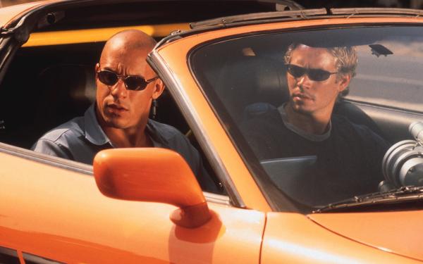 Vin deisel and Paul Walker wear sunglasses sitting in an orange sports car with the hood down